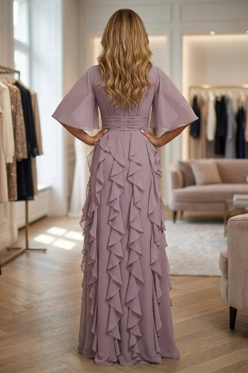 A woman stands with her back to the camera wearing a floor-length mauve dress with wide flutter sleeves, a fitted waist, and cascading ruffles down the skirt, photographed in a boutique interior.