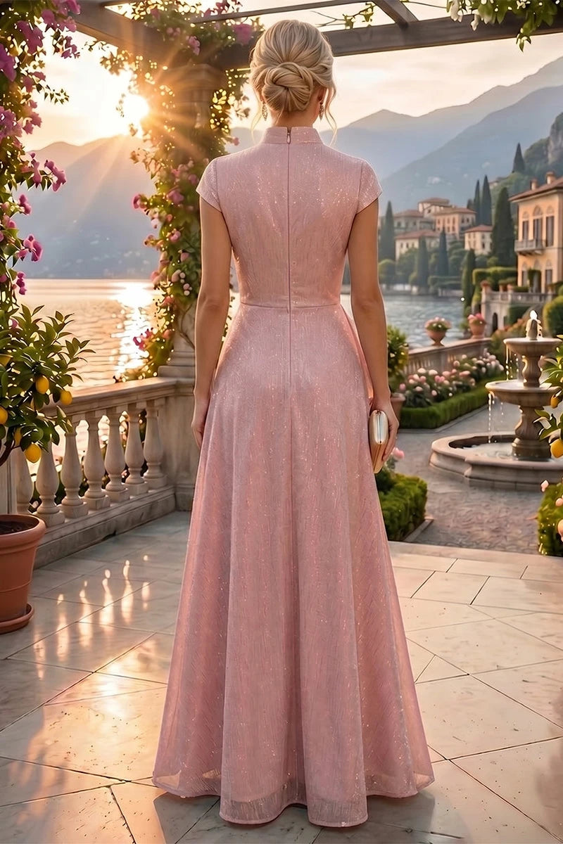 A woman stands on a sunlit terrace overlooking a lake and mountains, wearing a floor-length blush pink gown with cap sleeves and a high neckline, shown from the back.