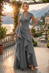 A woman stands on a sunlit terrace overlooking a lake and mountains, wearing a floor-length gray wrap-style gown with cap sleeves, a draped waist, and layered ruffles. She is holding a small beige clutch and wearing gold-toned heels and jewelry.