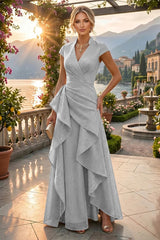 A woman stands on a sunlit terrace wearing a light silver-gray wrap-style maxi dress with cap sleeves and an asymmetrical draped skirt. The background shows a scenic lake, mountains, buildings, and garden plants at sunset.