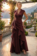A woman models a floor-length burgundy wrap gown with cap sleeves and a dramatic layered ruffled skirt, standing on a sunlit terrace overlooking a scenic lake and mountains.