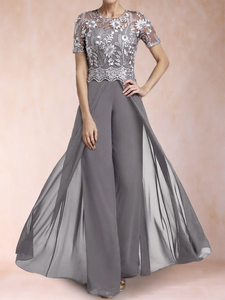 A woman modeling a floor-length gray gown with a sheer floral lace bodice, short sleeves, and a flowing draped skirt against a soft peach studio backdrop.