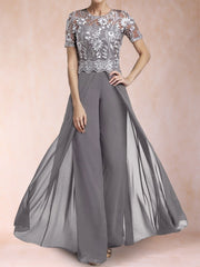 A woman modeling a floor-length gray gown with a sheer floral lace bodice, short sleeves, and a flowing draped skirt against a soft peach studio backdrop.