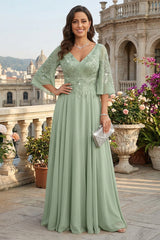 A woman stands on a sunlit terrace wearing a floor-length sage green gown with lace embroidery, three-quarter sleeves, and a V-neckline. She holds a small silver clutch in front of ornate stone planters and classical architecture.
