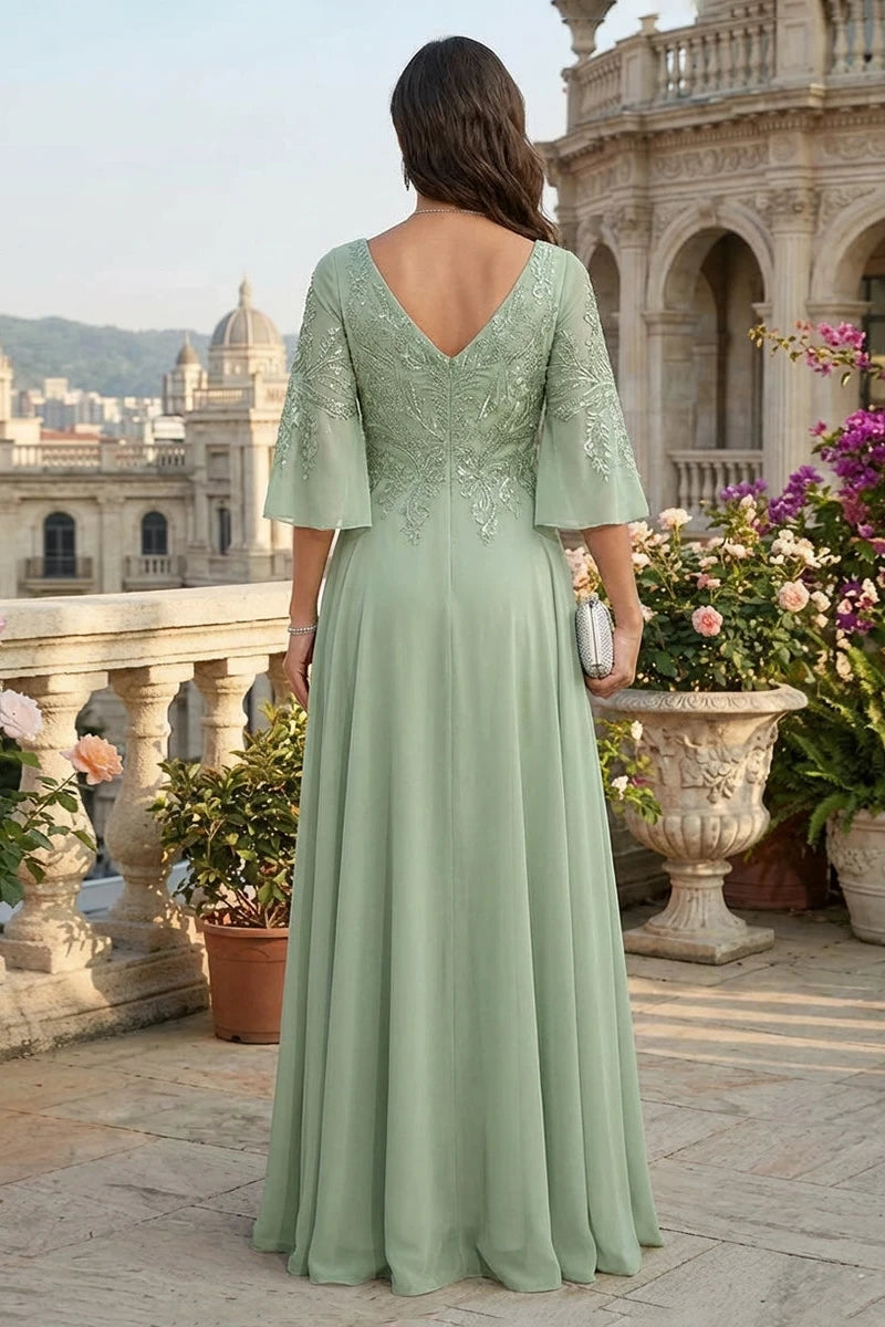 A woman stands on a sunlit terrace wearing a floor-length sage green gown with a deep V back, short flutter sleeves, and floral lace embroidery across the bodice. The background features a grand classical building, stone railings, and potted flowers.