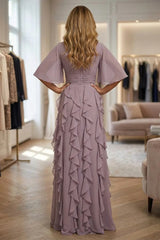 A woman stands with her back to the camera wearing a floor-length mauve dress with wide flutter sleeves, a fitted waist, and cascading ruffles down the skirt, photographed in a boutique interior.