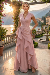 A woman models a floor-length blush pink gown with cap sleeves, a wrap-style V-neck bodice, and layered ruffle draping on a sunlit terrace overlooking a scenic lake and mountains.