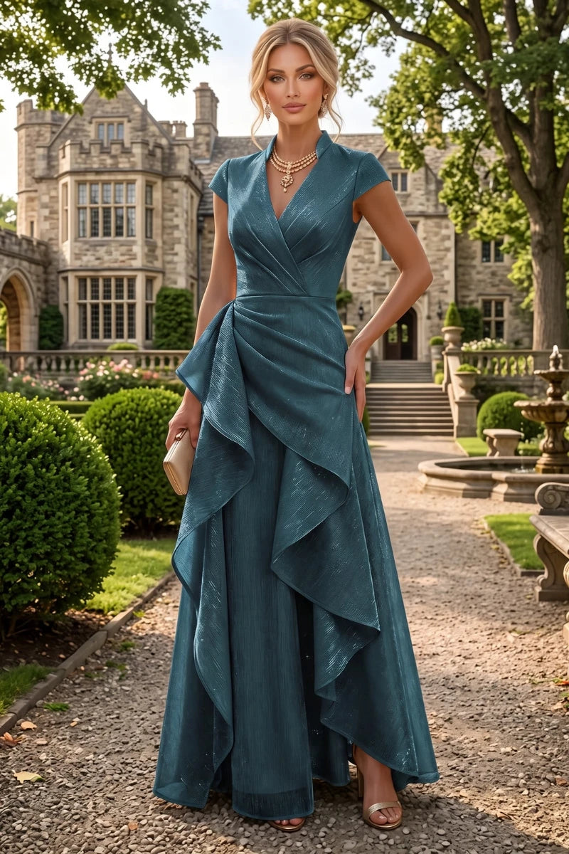 A woman stands on a gravel path in front of a large stone mansion, wearing a floor-length teal wrap-style gown with cap sleeves, a draped waist, and an asymmetrical layered hem.