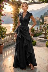 A woman models a floor-length black wrap gown with cap sleeves and an asymmetrical draped skirt, standing on a sunlit terrace with a lake, mountains, and buildings in the background.