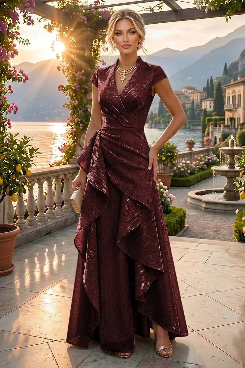 A woman models a floor-length burgundy wrap gown with cap sleeves and a dramatic layered ruffled skirt, standing on a sunlit terrace overlooking a scenic lake and mountains.