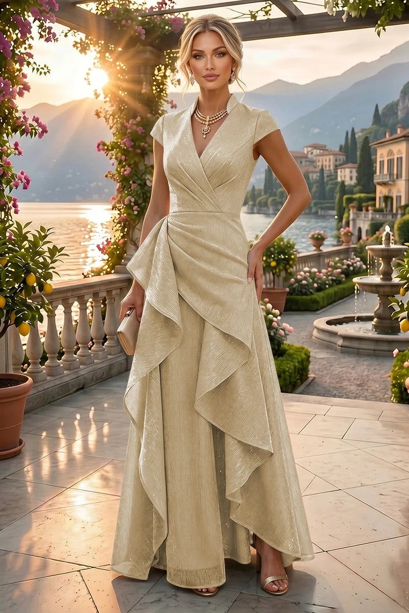 A woman stands on a sunlit terrace by a lake wearing a floor-length champagne gown with cap sleeves, a wrap-style bodice, and an asymmetrical draped skirt. The background shows mountains, water, flowers, and buildings at sunset.