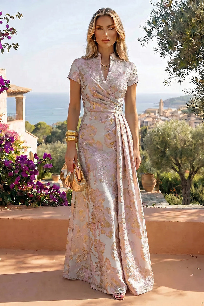A woman stands outdoors on a sunlit terrace wearing a floor-length draped gown with short sleeves and a gold-toned floral jacquard pattern. The dress has a high neckline and a self-tie waist.