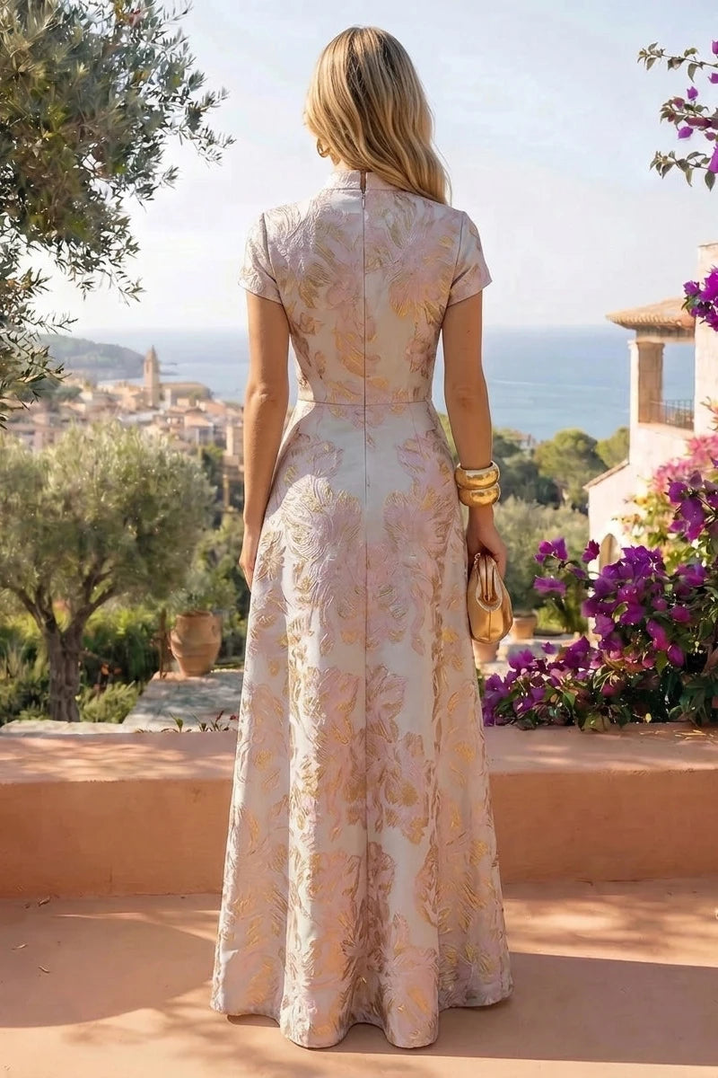 A woman stands outdoors on a terrace wearing a floor-length pale blush pink gown with a gold floral jacquard pattern. The dress has short sleeves, a fitted waist, and a high neckline.